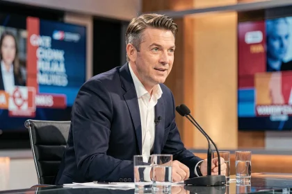 Animateur de débat politique assis au bureau d'un studio télévisé, dans un blazer marine, microphone devant lui, expression concentrée